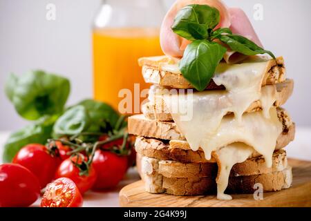 Vorrat an gerösteten, geschmolzenen, Käse gepressten Sandwiches mit Schinkenfleisch, Kirschtomaten, Orangensaft und Basilikumblättern auf Holzschneidebrett. Nahaufnahme Stockfoto