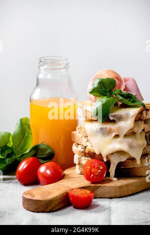 Vorrat an gerösteten, geschmolzenen, Käse gepressten Sandwiches mit Schinkenfleisch, Kirschtomaten, Orangensaft und Basilikumblättern auf Holzschneidebrett auf weißem m Stockfoto