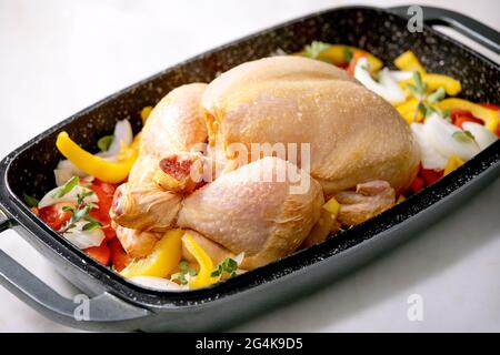 Rohes organisches, ungekochtetes Hähnchen mit geschnittenem Gemüse, Paprika, Zwiebeln und Kräutern im Ofenblech, fertig zum Kochen. Weißer Marmortisch. Stockfoto