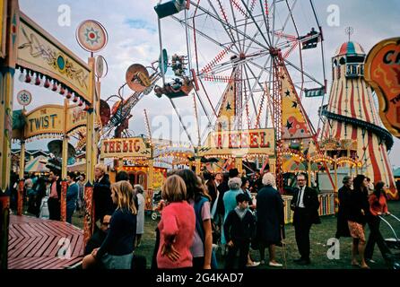 Menschenmassen in der Abenddämmerung auf der Bridgewater Fair, Bridgewater, Somerset, England, Großbritannien im September 1971. Die Fahrten umfassen das große Rad, Helter-Skelter und verschiedene Kreisverkehre und Karussells. Die Lichter sind an und traditionelle, bunte Zeichen von handbemalten Fairground-Schriftzügen und Kunst sind sichtbar. Die St. Matthews Messe stammt aus dem Jahr 1249. Die traditionelle, jährlich stattfindende viertägige Veranstaltung findet im September in St. Matthew’s Field statt – ein altes Foto aus den 1970er Jahren. Stockfoto