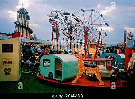 Menschenmassen in der Abenddämmerung auf der Bridgewater Fair, Bridgewater, Somerset, England, Großbritannien im September 1971. Die Fahrten umfassen das große Rad, Helter-Skelter und verschiedene Kreisverkehre und Karussells. Die Lichter sind an und traditionelle, bunte Zeichen von handbemalten Fairground-Schriftzügen und Kunst sind sichtbar. Die St. Matthews Messe stammt aus dem Jahr 1249. Die traditionelle, jährlich stattfindende viertägige Veranstaltung findet im September in St. Matthew’s Field statt – ein altes Foto aus den 1970er Jahren. Stockfoto