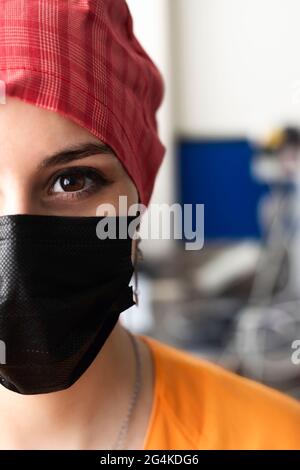 Nahaufnahme der Hälfte des Gesichts eines attraktiven jungen Arztes, der eine schwarze OP-Maske, eine rote OP-Kappe und eine orangefarbene Uniform trägt und bereit ist, einzutreten Stockfoto