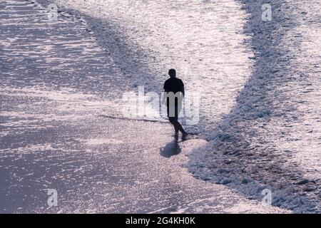 Ein Mann, der durch das Abendlicht geschildet wurde und aus dem Meer ging, folgte kleinen Wellen, die sanft am Fistral Beach in Newquay in Cornwall flossen. Stockfoto