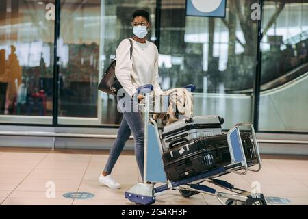 Weibliche Reisende am Flughafenterminal mit Gepäckwagen während einer Pandemie. Afrikanische junge Frau in Gesichtsmaske am Flughafen. Stockfoto