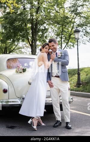 Glückliche Frau im Hochzeitskleid umarmen Bräutigam in der Nähe Auto auf der Straße Stockfoto