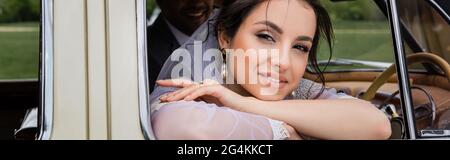 Lächelnde Braut im Schleier Blick auf die Kamera in der Nähe verschwommener Bräutigam im Oldtimer, Banner Stockfoto