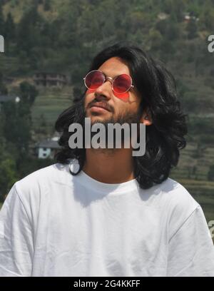 Porträt eines gut aussehenden, langhaarigen indischen jungen Mannes, der eine Sonnenscheibe trägt und nach oben schaut, eines hübschen bärtigen Mannes, der draußen mit einem weißen T-Shirt steht Stockfoto