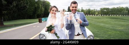 Fröhlicher Bräutigam zeigt auf Ring neben der Braut mit Blumenstrauß und Oldtimer, Banner Stockfoto