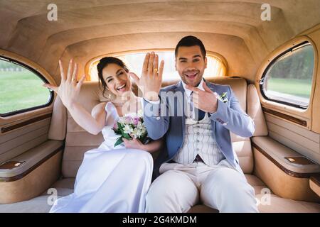 Fröhlicher Bräutigam zeigt auf Ring auf Finger in der Nähe der Braut im Retro-Auto Stockfoto