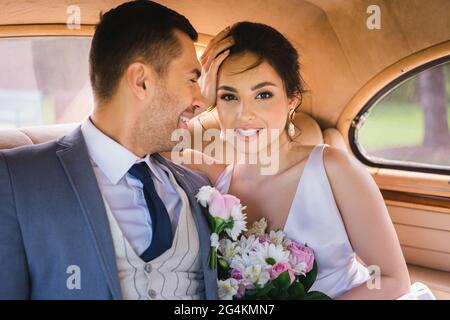 Positive Braut mit Blumenstrauß Blick auf Kamera in der Nähe Bräutigam im Retro-Auto Stockfoto