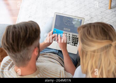 Online-Shopping von zu Hause aus. Ein hübscher Mann hält eine Bankkarte in der Hand, während ihre Frau neben ihm sitzt und einen Laptop benutzt, während sie online Geld sendet. Stockfoto