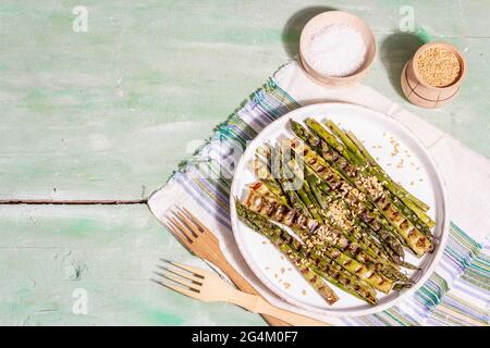 Gereifter gegrillter Spargel. Goldene Leinsamen, Meersalz, gesundes und gesundes Lebensmittelkonzept. Moderner, harter, dunkler Schatten. Hellgrüner Tisch, Draufsicht Stockfoto