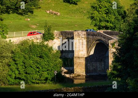 Landschaftlich reizvolle Aussicht auf die historische, steinerne, gewölbte Brücke über den Fluss Wharfe (2 Autos überqueren die Brücke) - Barden Bridge, Yorkshire Dales, England, Großbritannien. Stockfoto