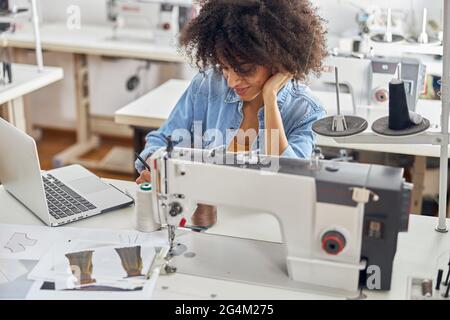 Die afroamerikanische Designerin zeichnet mit Nähmaschine und Laptop Skizzen am Tisch Stockfoto