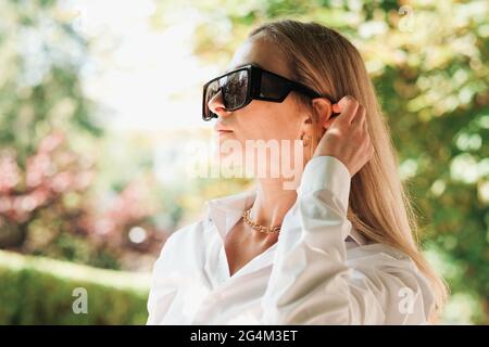 Nahaufnahme Porträt einer selbstbewussten Frau, die in der Stadt unterwegs ist, einer Unternehmerin, die eine Sonnenbrille trägt und ihr Haar hinter das Ohr nimmt. Stockfoto