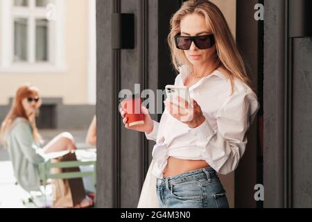 Elegante Frau, die aus einem Café kommt, Tasse Kaffee in der Hand hält, während sie im Smartphone schaut, selbstbewusste Unternehmerin, die eine Sonnenbrille trägt und Stockfoto