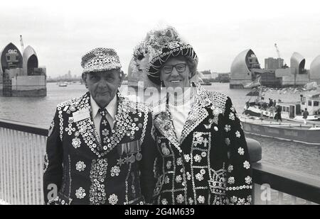 Die historischen 1980er Jahre, Londons Pearly King und Queen stehen für ihr Bild auf der Südseite der Themse in New Charlton, Greenwich, London, England, Großbritannien. Vertreter der Londoner Arbeiterkultur, der Pearlies, wie sie aufgrund ihrer mit Perlmutt-Knöpfen verzierten Kleidung bekannt sind, stehen vor der neu errichteten Thames Water Barrier, einem einziehbaren Hochwasserschutzsystem. Seit 1982 in Betrieb, wurde es 1984 von Ihrer Majestät Königin Elizabeth II. Offiziell eröffnet. Die Barriere schützt das Zentrum Londons vor einem Sturmflut. Stockfoto