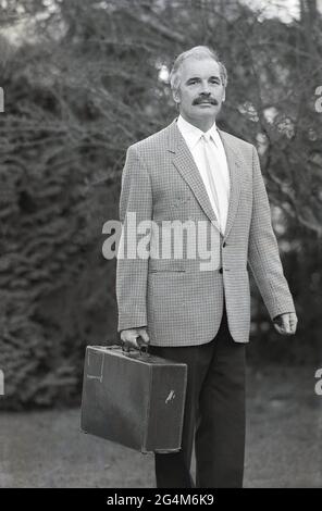 In den 1980er Jahren, ein Geschäftsmann in einer Jacke mit Karokmuster und Krawatte, der draußen mit einer alten Aktentasche aus Leder stand, England, Großbritannien. Eine Aktentasche, die zum Tragen von Papieren und Akten verwendet wird und mit einem Griff ausgestattet ist, stammt von den Taschen oder Umhängetaschen ab, die im 14. Jahrhundert zum Tragen von Geld und Wertsachen verwendet wurden. Der Name stammt von den Taschen oder Fällen, in denen Anwälte ihre juristischen Papiere, ihre "Briefs", die sie dem Gericht vorstellten, mit sich brachten. Es wird vermutet, dass die so genannte erste moderne rechteckige Aktentasche um die 1850er Jahre erfunden wurde. Stockfoto