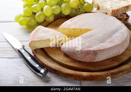 Reblochon auf Schneidebrett mit Trauben und Brotscheiben auf weißem Holztisch. Französischer Käse, der in der Alpenregion Savoyen aus rohem c Stockfoto