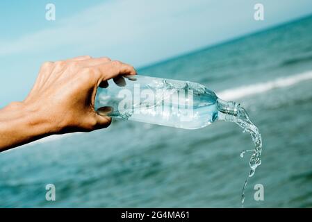 Ein junger Kaukasusmann gießt Wasser aus einer wiederverwendbaren Glasflasche vor dem Ozean Stockfoto