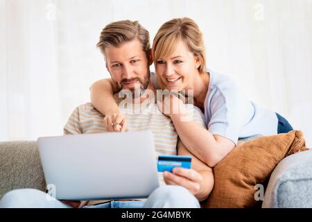 Online-Shopping von zu Hause aus. Ein hübscher Mann hält eine Bankkarte in der Hand, während ihre Frau neben ihm sitzt und einen Laptop benutzt, während sie online einkaufen. Stockfoto