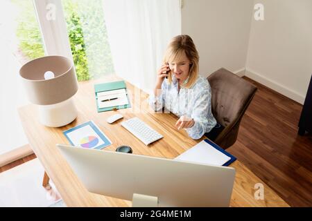 Geschäftsfrau, die einen Anruf hat, während sie am Schreibtisch hinter einem Computer sitzt. Heimbüro. Stockfoto