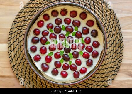 Die Nahaufnahme des hausgemachten Käsekuchen wird mit frischen Kirschen und einem Zweig Minze in einer runden Kuchenform auf einem Holztisch gekrönt Stockfoto