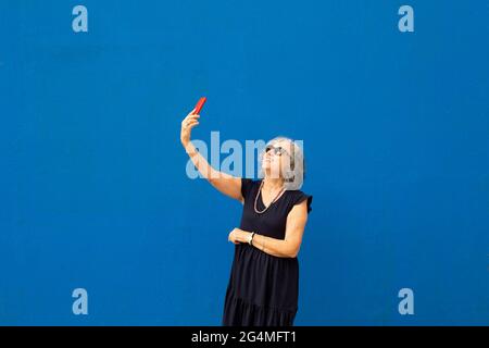 Ältere grauhaarige Frau, die ein Selfie mit einem roten Smartphone gegen eine blaue Wand nimmt Stockfoto