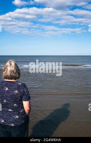 Eine blonde Frau genießt es, das Meer zu beobachten, während die Wellen an einem schönen sonnigen Tag in Großbritannien zum Strand Rollen Stockfoto