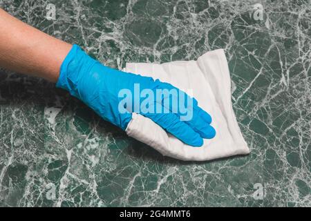 Die Hand einer Frau in einem blauen Haushaltshandschuh wischt eine weiße Servietten-Marmoroberfläche in der Küche aus nächster Nähe ab. Stockfoto