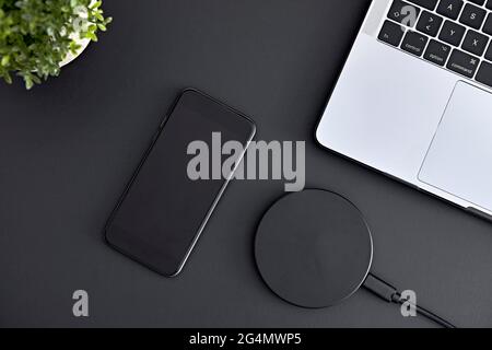 Blick auf Smartphone, Laptop und kabelloses Ladegerät, die von direkt oben auf dem grauen Schreibtisch liegen. Moderne Technologie, drahtlose Geräte und Übertragung von en Stockfoto