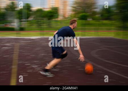 Junger Rotschopf, der tagsüber auf einem Sportplatz auf der Straße mit hoher Geschwindigkeit einen Basketball läuft und dribbt, verschwommen die Bewegung Stockfoto