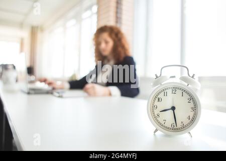 Schöne junge rothaarige Frau in einem Business-Anzug arbeitet in einem Büro mit einem Laptop, Wecker im Vordergrund Stockfoto