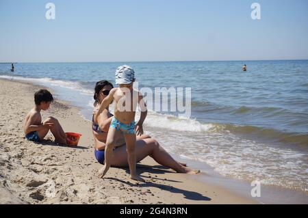 POBIEROWO, POLEN - 22. Jun 2021: Frau, die an einem warmen Tag mit zwei Jungen auf Sand in der Nähe des Wassers am Strand sitzt. Stockfoto