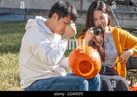 Bloomington USA 10-19-2019 ein Paar sitzt draußen auf dem Campus und schnitzt Kürbis-Jack-o-Laterne mit einem Mädchen, das ihn bei seiner Arbeit videogeführt hat Stockfoto