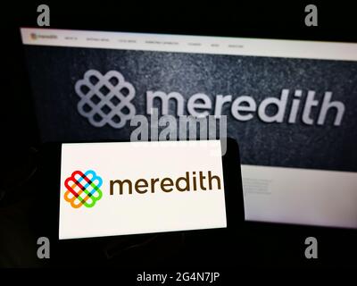Person, die Mobiltelefon mit dem Logo des amerikanischen Medienunternehmens Meredith Corporation auf dem Bildschirm vor der Business-Webseite hält. Konzentrieren Sie sich auf die Telefonanzeige. Stockfoto