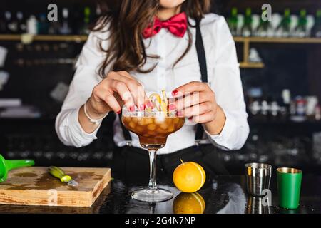 Eine nicht erkennbare Barkeeperin in stilvollem Outfit, die einen Cocktail mit Zitronenschale schmückt, während sie an der Theke in der modernen Bar steht Stockfoto