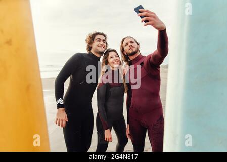 Eine Gruppe glücklicher Surfer-Freunde, die in Neoprenanzügen gekleidet sind und in der Nähe von Surfbrettern stehen, während sie während des Trainings Selfie mit dem Smartphone am Strand machen Stockfoto