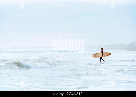 Seitenansicht des in Neoprenanzug gekleideten Surfers, der mit dem Surfbrett zum Wasser hinausschaute, um während des Sonnenaufgangs eine Welle am Strand zu fangen Stockfoto
