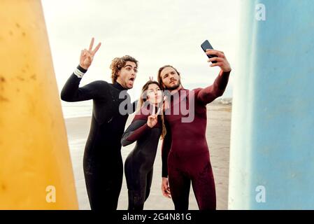 Eine Gruppe glücklicher Surfer-Freunde, die in Neoprenanzügen gekleidet sind und in der Nähe von Surfbrettern stehen, während sie während des Trainings Selfie mit dem Smartphone am Strand machen Stockfoto