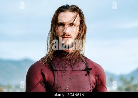 Junger, hübscher Surfer mit langen Haaren, die in Neoprenanzug gekleidet sind und während des Sonnenaufgangs auf den Strand blicken Stockfoto