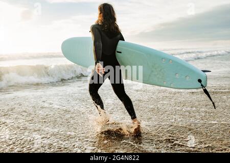 Seitenansicht des in Neoprenanzug gekleideten Surfers, der mit dem Surfbrett zum Wasser läuft, um während des Sonnenaufgangs eine Welle am Strand zu fangen Stockfoto