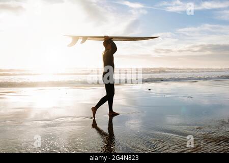 Seitenansicht des in Neoprenanzug gekleideten Surfers, der am Morgen mit dem Surfbrett über seinem Kopf am Strand spazierenging, mit Sonnenaufgang im Hintergrund Stockfoto
