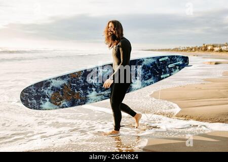 Seitenansicht des in Neoprenanzug gekleideten Surfers, der mit dem Surfbrett zum Wasser läuft, um während des Sonnenaufgangs eine Welle am Strand zu fangen Stockfoto