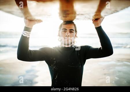 Porträt eines jungen Surfers, der in einem Neoprenanzug gekleidet ist und während des Sonnenaufgangs die Kamera am Strand mit dem Surfbrett über dem Kopf anschaut Stockfoto