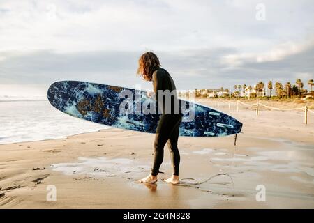 Seitenansicht des in Neoprenanzug gekleideten Surfers, der mit dem Surfbrett zum Wasser läuft, um während des Sonnenaufgangs eine Welle am Strand zu fangen Stockfoto