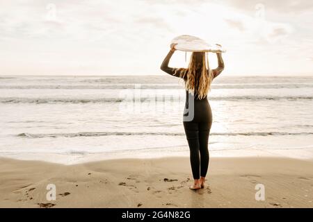 Rückansicht einer nicht erkennbaren weiblichen Surferin, die in einem Neoprenanzug gekleidet ist und im Hintergrund beim Sonnenaufgang das Surfbrett am Strand hält Stockfoto