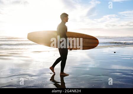 Seitenansicht des in Neoprenanzug gekleideten Surfers, der am Morgen mit dem Surfbrett am Strand spazierenging, während der Sonnenaufgang im Hintergrund zu beobachten war Stockfoto