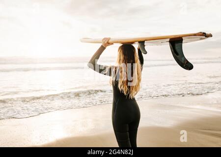 Rückansicht einer nicht erkennbaren weiblichen Surferin, die in einem Neoprenanzug gekleidet ist und im Hintergrund beim Sonnenaufgang das Surfbrett am Strand hält Stockfoto
