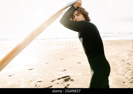 Seitenansicht eines lächelnden Surfers in Neoprenanzug, der während des Sonnenaufgangs im Hintergrund die Kamera mit dem Surfbrett am Strand betrachte Stockfoto
